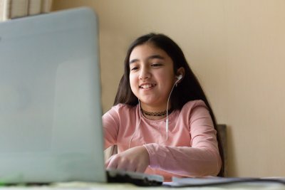 Young girl sitting at the dining table with laptop at home schooling, online virtual classroom video conference, distant education. Active participation at the lesson with earphones, doing homework.