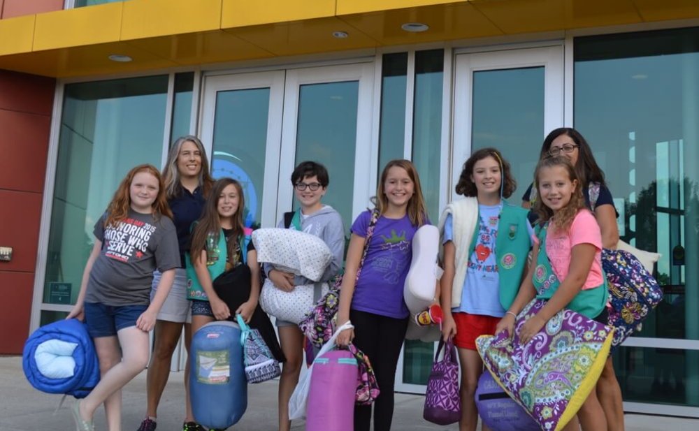 A group of girls pose in front of the science center with sleeping bags and pillows for their overnight camp-in